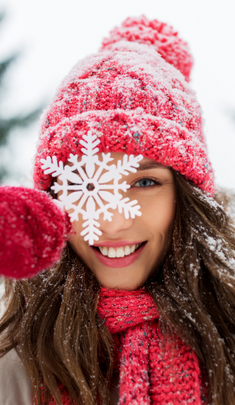 Smiling person in red winter hat and scarf holds a large white snowflake in front of one eye. Snow lightly dusts their clothing, creating a playful, joyful atmosphere.